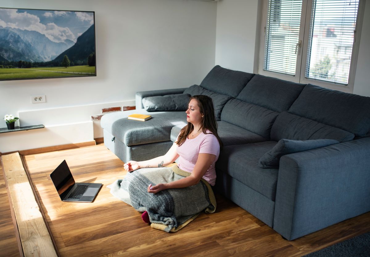 a woman sits on the floor of her living room with a blanket as she follows a guided meditation class online on her laptop.
